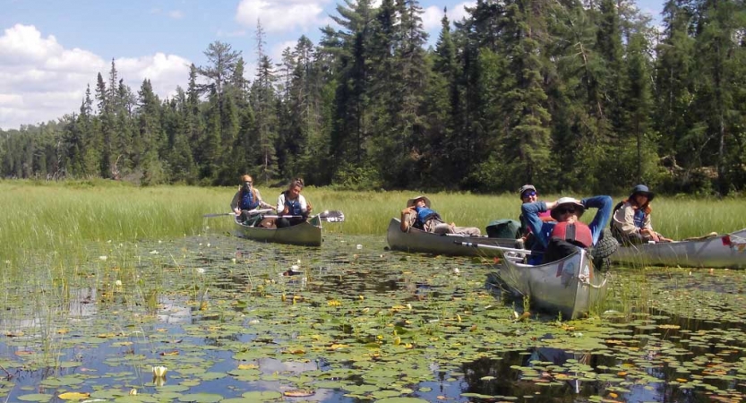 a group of teens rest in the canoes on calm water. The water is covered with lily pads, and there is a line of evergreen trees in the background. 